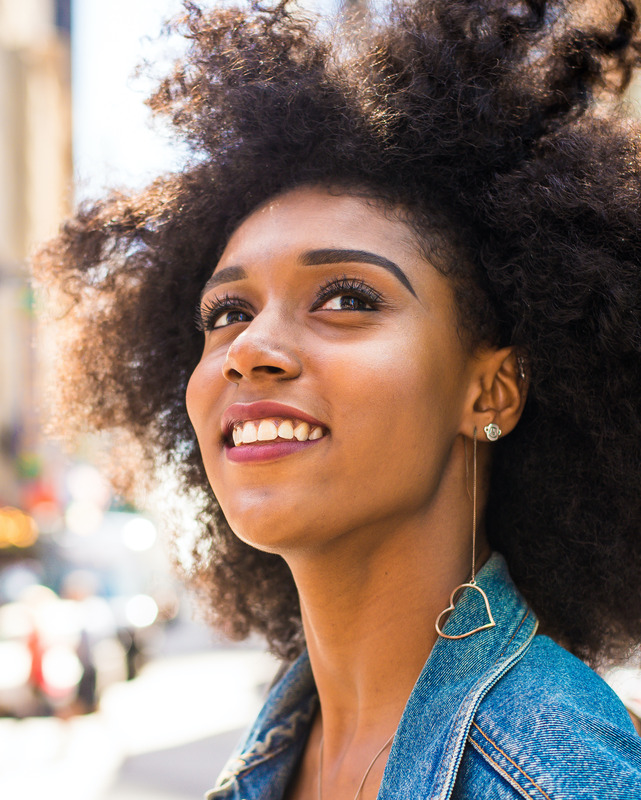 Photo of a woman with great skin outside in a city. She is wearing long heart earrings.