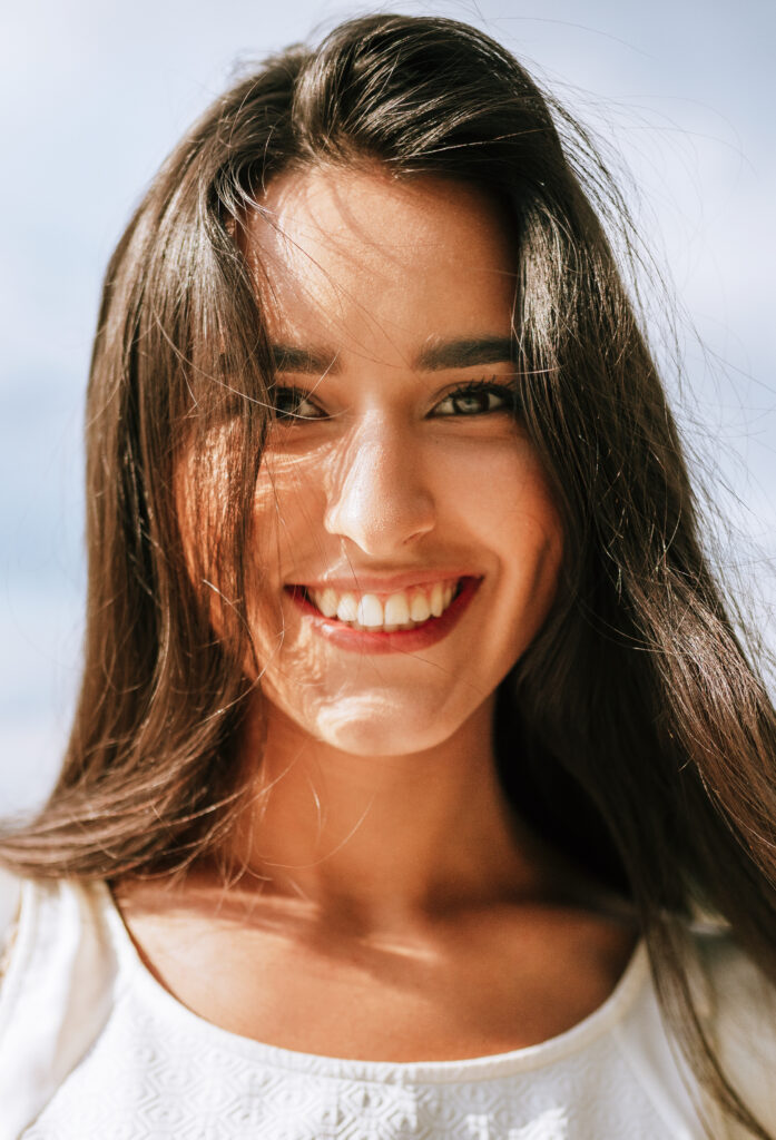 Happy beautiful young woman smiling broadly with healthy toothy smile and with a windy blowing long hair during walking in the park, posing on nature background.